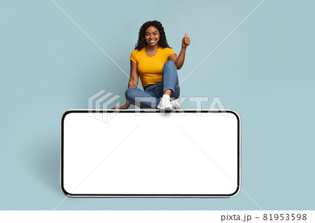 Cheerful african american young woman sitting on huge cellphone, mockup 81953598
