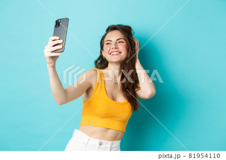 Beautiful modern girl taking selfies on vacation, posing in summer clothes and looking at smartphone camera, standing against blue background Beautiful modern girl taking selfies on vacation, posing in summer clothes and looking at smartphone camera, standing against blue background 81954110