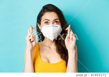 Health, covid-19 and lockdown concept. Close up of hopeful young woman in respirator mask, cross fingers and making wish, praying and looking up, blue background Health, covid-19 and lockdown concept. Close up of hopeful young woman in respirator mask, cross fingers and making wish, praying and looking up, blue background 81954172