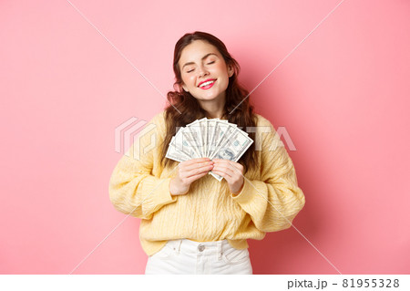 Attractive young woman with pleased smile, hugging her money with satisfied expression, dreaming of shopping, standing against pink background Attractive young woman with pleased smile, hugging her money with satisfied expression, dreaming of shopping, standing against pink background 81955328