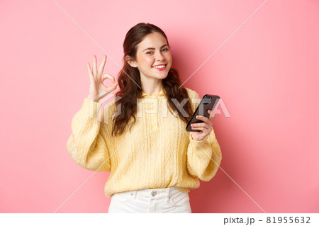 Technology and online shopping. Young female model shows okay sign and use mobile phone app, praise good shop, say yes, stands against pink background Technology and online shopping. Young female model shows okay sign and use mobile phone app, praise good shop, say yes, stands against pink background 81955632