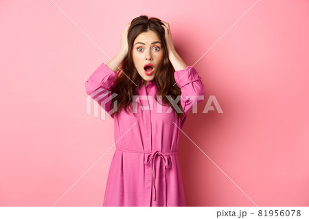 Shocked glamour girl gasping and looking startled, holding hands on head, standing in panic against pink background 81956078