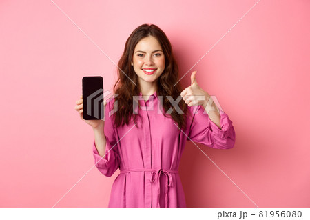 Technology concept. Young girl in dress, shows her smartphone screen and thumbs up, nod in approval, praise good app, standing over pink background 81956080