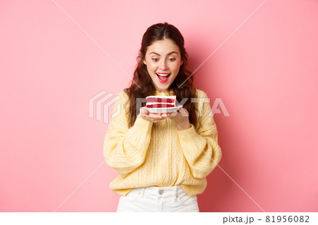 Celebration and holidays. Happy birthday girl stares at delicious bday cake dessert and smiles, stands against pink background Celebration and holidays. Happy birthday girl stares at delicious bday cake dessert and smiles, stands against pink background 81956082