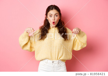 Wow super cool. Excited brunette girl pointing fingers down, gasping wondered, showing direction way, indicating at bottom logo, standing over pink background Wow super cool. Excited brunette girl pointing fingers down, gasping wondered, showing direction way, indicating at bottom logo, standing over pink background 81956421