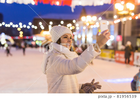 Christmas, winter, technology and leisure concept - happy young woman taking picture with smartphone on ice skating rink outdoors. 81956539