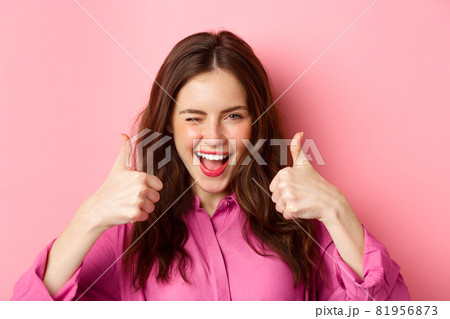 Cheeky glamour girl winking at you and shows thumbs up, praise great work, good choice, standing over pink background Cheeky glamour girl winking at you and shows thumbs up, praise great work, good choice, standing over pink background 81956873
