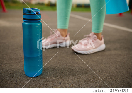 Close-up of a blue plastic bottle of water on the asphalt of sports ground next to sporty woman's feet. Health and body care concept Close-up of a blue plastic bottle of water on the asphalt of sports ground next to sporty woman's feet. Health and body care concept 81958891