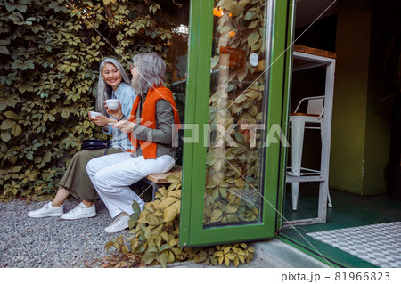 Cheerful senior ladies friends with cups of drinks sit on bench in cafe yard 81966823