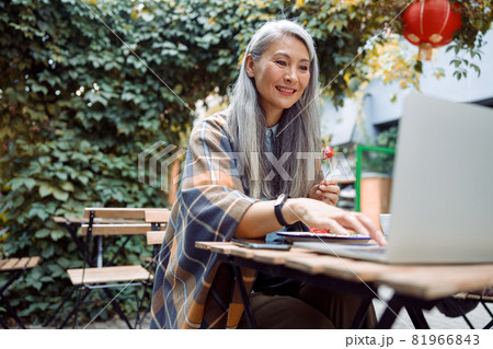 Smiling mature Asian woman eats strawberry dessert working on laptop at table outdoors Smiling mature Asian woman eats strawberry dessert working on laptop at table outdoors 81966843