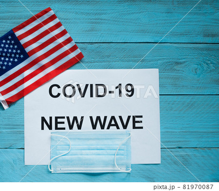 On a blue wooden background is a sheet with the inscription: covid -19 new wave, a medical mask and a U.S. flag. Concept of the next wave of covid infections in the United States On a blue wooden background is a sheet with the inscription: covid -19 new wave, a medical mask and a U.S. flag. Concept of the next wave of covid infections in the United States 81970087