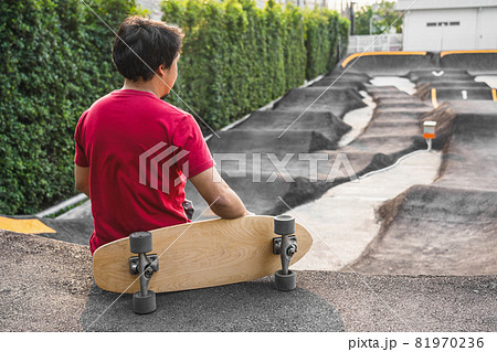 Asian man sitting with surfskate or skate board in pumptrack skate Park when sunrise time over photo blur of pumptrack curve, extream sport, healthy and exercise, fashion in covid19 concept 81970236