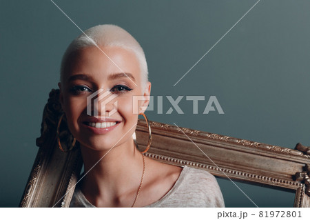 Millenial young woman with short blonde hair holds gilded picture frame in hands behind her face portrait. 81972801