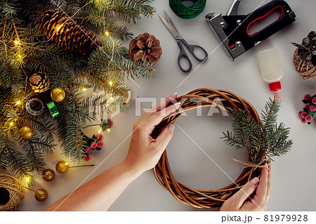 Female hands decorate the Christmas wreath with spruce branches with a red berry and forest cones. White background. New Year's concept. Female hands decorate the Christmas wreath with spruce branches with a red berry and forest cones. White background. New Year's concept. 81979928