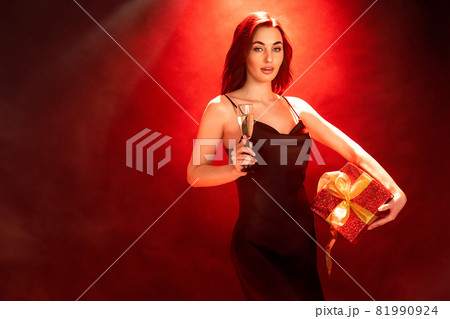 Woman in black dress holds the gift red box and glass of champagne. Party time. Holiday concept Woman in black dress holds the gift red box and glass of champagne. Party time. Holiday concept 81990924