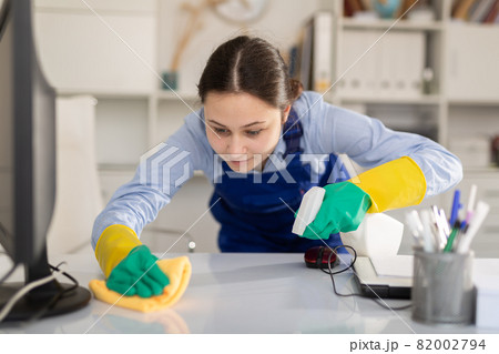 Cleaning lady in overalls wipes dust from table in office Cleaning lady in overalls wipes dust from table in office 82002794