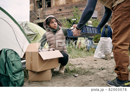 Dark-skinned teenage girl crouching at stuff box and putting tin can into basket while refugees collecting food for dinner in camp Dark-skinned teenage girl crouching at stuff box and putting tin can into basket while refugees collecting food for dinner in camp 82003078