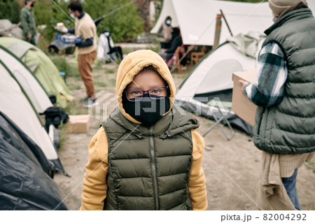 Portrait of mixed race child in hoodie, vest, eyeglasses and cloth mask standing in refugee camp 82004292