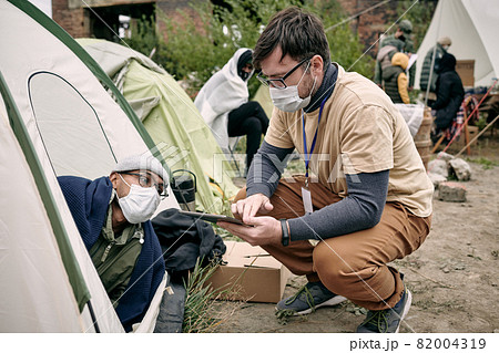 Young social worker with badge crouching at tent and making notes in tablet while talking to refugee Young social worker with badge crouching at tent and making notes in tablet while talking to refugee 82004319