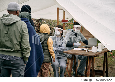 Medical specialists in protective masks sitting at table in tent and checking health of migrants standing in line Medical specialists in protective masks sitting at table in tent and checking health of migrants standing in line 82005422
