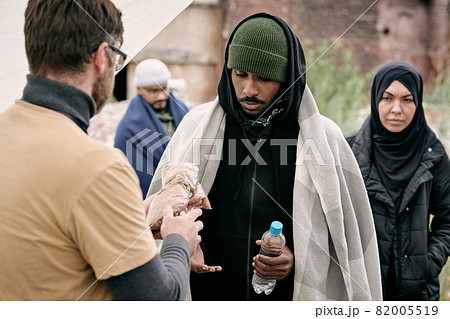 Social worker giving water and cereal to black man under plaid while providing food to refugees outdoors 82005519