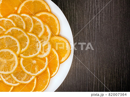 Sliced orange and lemon in a white plate on a dark wooden texture background, copy space 82007364