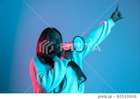One African beautiful woman with long straight hair shouting at megaphone isolated on blue studio background in neon light. Concept of human emotions One African beautiful woman with long straight hair shouting at megaphone isolated on blue studio background in neon light. Concept of human emotions 82010939
