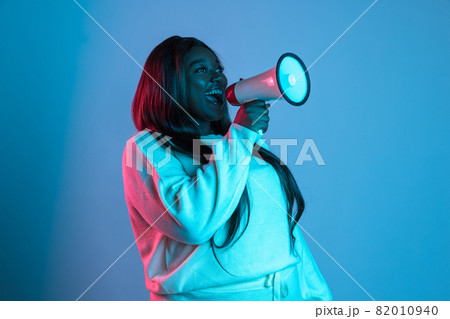 One African beautiful woman with long straight hair shouting at megaphone isolated on blue studio background in neon light. Concept of human emotions One African beautiful woman with long straight hair shouting at megaphone isolated on blue studio background in neon light. Concept of human emotions 82010940