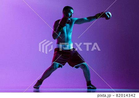 Side view full-length portrait of one professional boxer in red uniform training isolated over purple background in neon light. Straight punch move 82014342