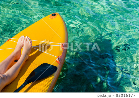 stand up paddle board with visible legs of a young girl and a paddle on the turquoise surface of the ocean 82016177