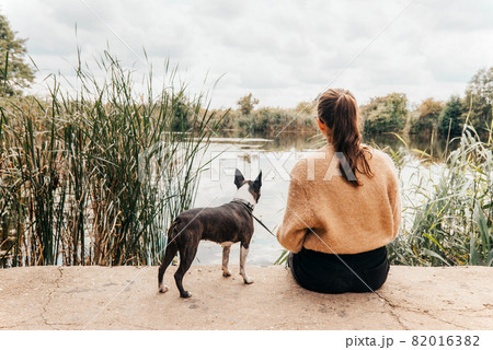 Teenage girl sitting with a Boston Terrier dog on the lakeshore - with her back turned, her dog staring at water - Autumn scenery Teenage girl sitting with a Boston Terrier dog on the lakeshore - with her back turned, her dog staring at water - Autumn scenery 82016382