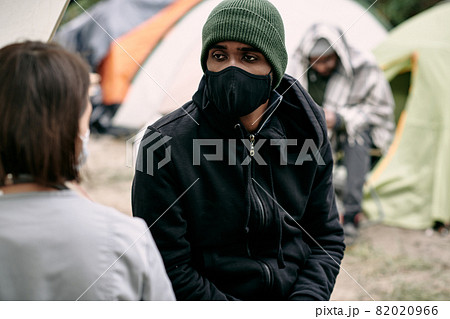Sick young Black refugee man in facial mask and hat talking to nurse in migrant camp Sick young Black refugee man in facial mask and hat talking to nurse in migrant camp 82020966