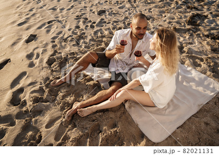 Portrait of a young couple sitting on the beach and drinking wine 82021131