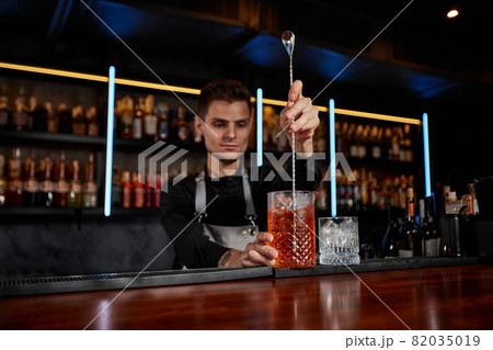 Barman stirring ice cubes in cocktail glass with spoon 82035019