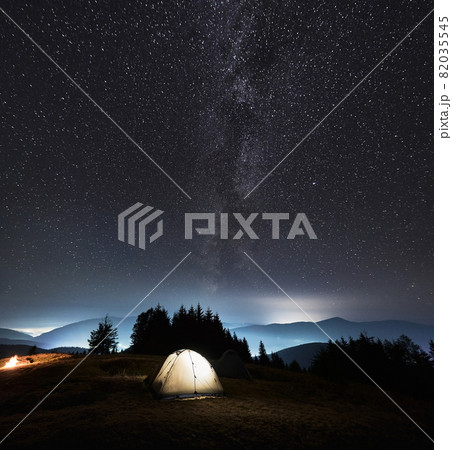 Fantastic view of night starry sky over grassy hill with illuminated tourist tent. Magnificent scenery of mountain valley with camp tent under night sky with stars and Milky Way. Concept of camping. 82035545