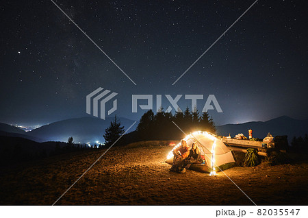 Couple travelers sitting inside illuminated camp tent and looking at evening sky with stars. Magnificent view of blue starry sky over mountain valley. Concept of travelling, hiking, night camping. 82035547