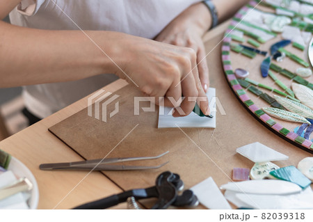 Workplace of the mosaic master: women's hands holding tool for mosaic details in the process of making a mosaic Workplace of the mosaic master: women's hands holding tool for mosaic details in the process of making a mosaic 82039318