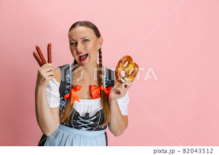 Adorable Oktoberfest woman, waitress wearing a traditional Bavarian or german dirndl holding bretzel isolated on pink studio background. 82043512