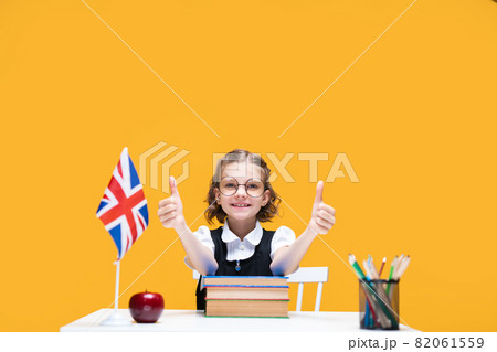 Happy caucasian schoolgirl sitting at the desk showing thumbs up. English lesson Great Britain flag Happy caucasian schoolgirl sitting at the desk showing thumbs up. English lesson Great Britain flag 82061559