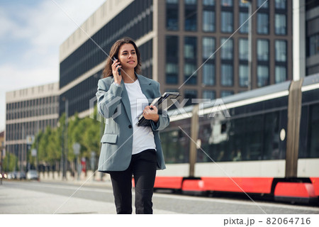 Beautiful formal dressed business woman in suit with a laptop and notebook outdoor. Brunette caucasian female, standing near office. 82064716