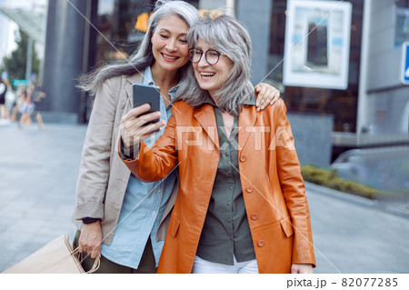 Smiling silver haired lady and companion with shopping bags take selfie on modern city street 82077285