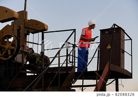 Male worker in protective helmet holding clipboard while monitoring work of petroleum pump jack. Petroleum engineer in work vest working in oil field. Concept of petroleum industry and oil extraction. Male worker in protective helmet holding clipboard while monitoring work of petroleum pump jack. Petroleum engineer in work vest working in oil field. Concept of petroleum industry and oil extraction. 82088326