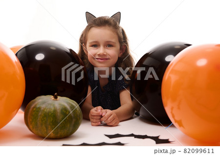 Smiling adorable child girl in carnival costume, little witch lying down on white background with copy space next to pumpkins, handmade bats and colorful orange black balloons. Halloween party concept 82099611