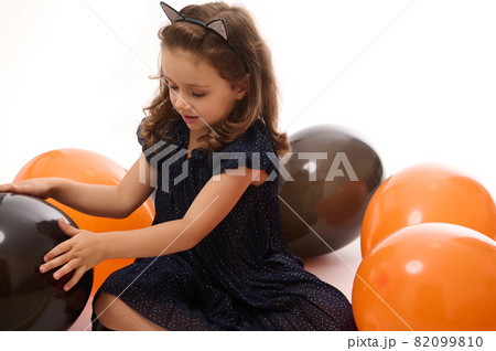 Adorable baby girl, 4 years old child wearing an evening dress and hoop with cat ears, having fun playing with bright orange and black balloons on white background with copy space. Halloween concept Adorable baby girl, 4 years old child wearing an evening dress and hoop with cat ears, having fun playing with bright orange and black balloons on white background with copy space. Halloween concept 82099810