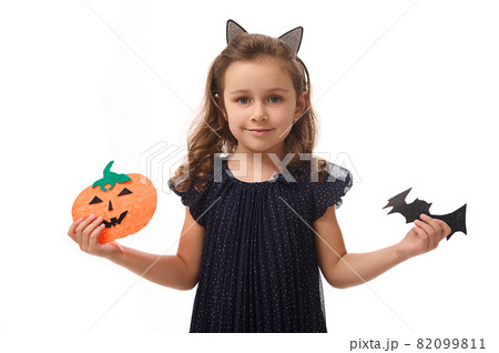 Pretty little girl with a hoop in the form of cat ears, dressed in a beautiful evening dress, holds felt-cut pumpkin and handmade bat, smiles looking at camera. Isolated on white background copy space Pretty little girl with a hoop in the form of cat ears, dressed in a beautiful evening dress, holds felt-cut pumpkin and handmade bat, smiles looking at camera. Isolated on white background copy space 82099811