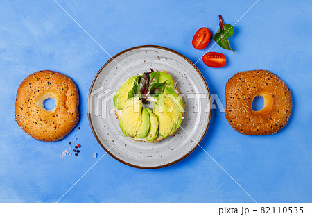 Bagel with avocado slices and salad leaves on a gray plate. On a blue background, horizontal 82110535