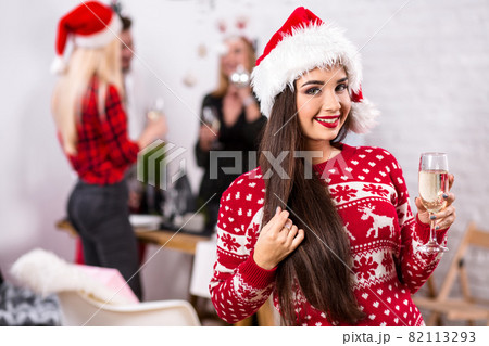 Portrait of a young woman with a glass of champagne at home on the foreground. Beautiful brunette in a Santa hat, red costume with deers Portrait of a young woman with a glass of champagne at home on the foreground. Beautiful brunette in a Santa hat, red costume with deers 82113293