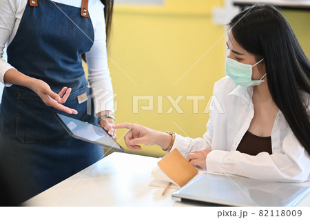 Young woman costumer wearing protective mask reading the menu on the tablet. Young woman costumer wearing protective mask reading the menu on the tablet. 82118009