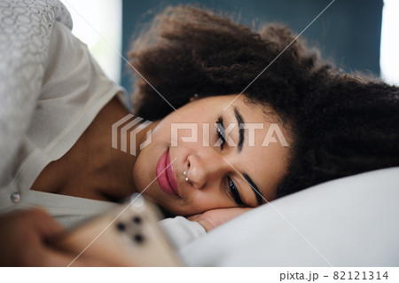 Front view portrait of young woman indoors on bed, using smartphone. 82121314