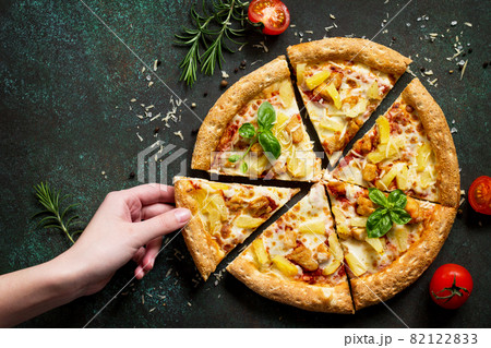 Pizza Hawaiian in a cardboard box on a dark stone or concrete background hands, taking pieces pizza. Copy space, top view flat lay background. Pizza Hawaiian in a cardboard box on a dark stone or concrete background hands, taking pieces pizza. Copy space, top view flat lay background. 82122833